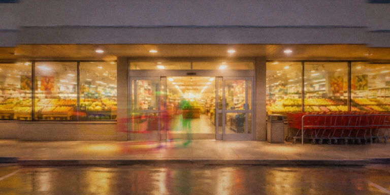 A lit grocery storefront at dusk, reflected in wet pavement